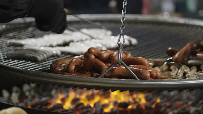 Chefs hands turn sausages on metal grill suspended on chains over fire at Christmas market in Germany in winter. Sausage barbecue of different types in evening at Christmas Fair. Bayerische Wurste. 