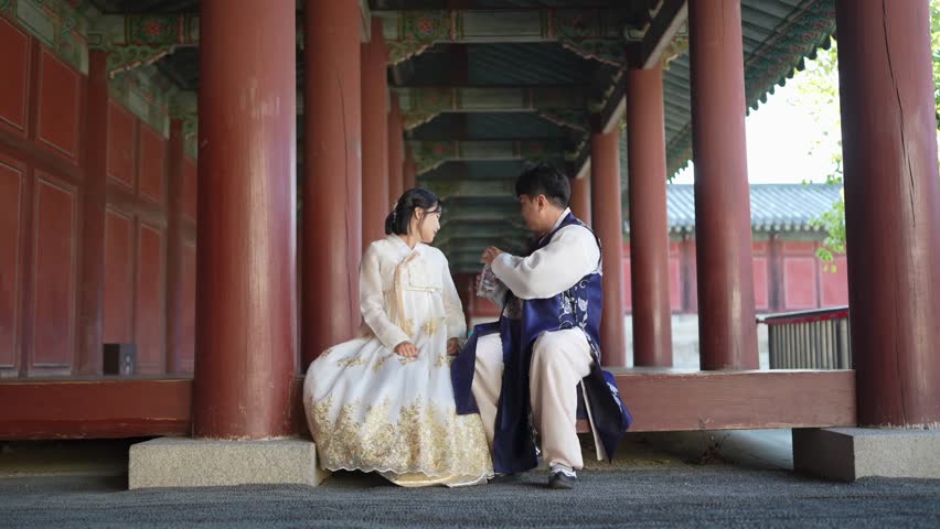 A Korean couple, a man in his 30s and a woman in her 20s, wearing hanbok from Seoul, South Korea, are sitting in a historical building, drinking water and using their smartphones. Slow motion video.