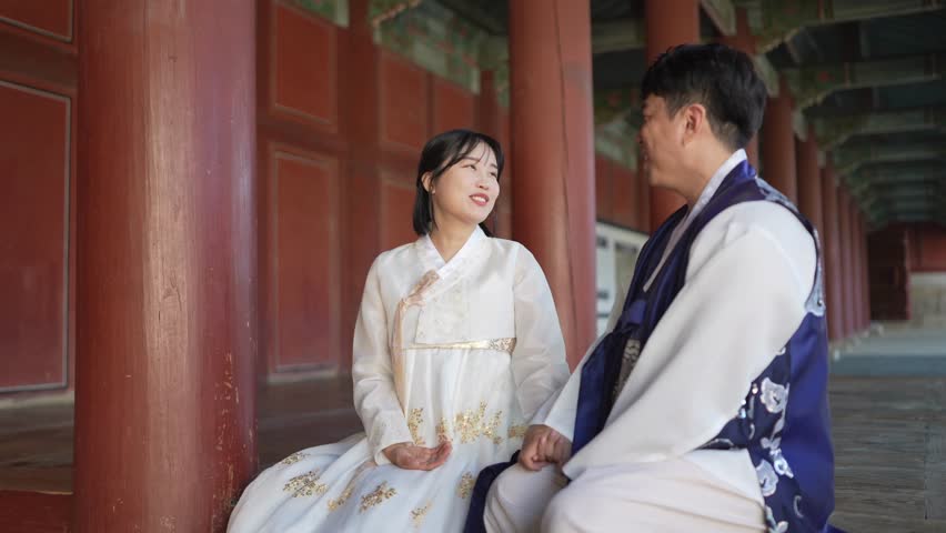 A Korean couple, a man in his 30s and a woman in her 20s, both from Seoul, South Korea, wearing Hanbok, are sitting side by side in a historical building and talking. Slow motion video.