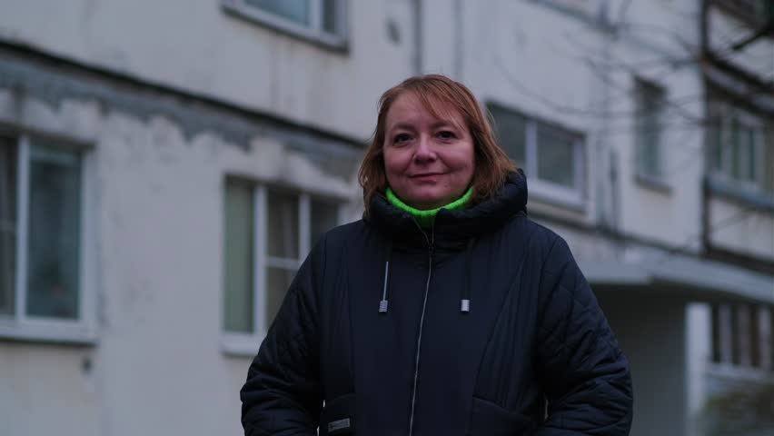 Woman in winter coat stands outside apartment building in urban setting during early evening hours