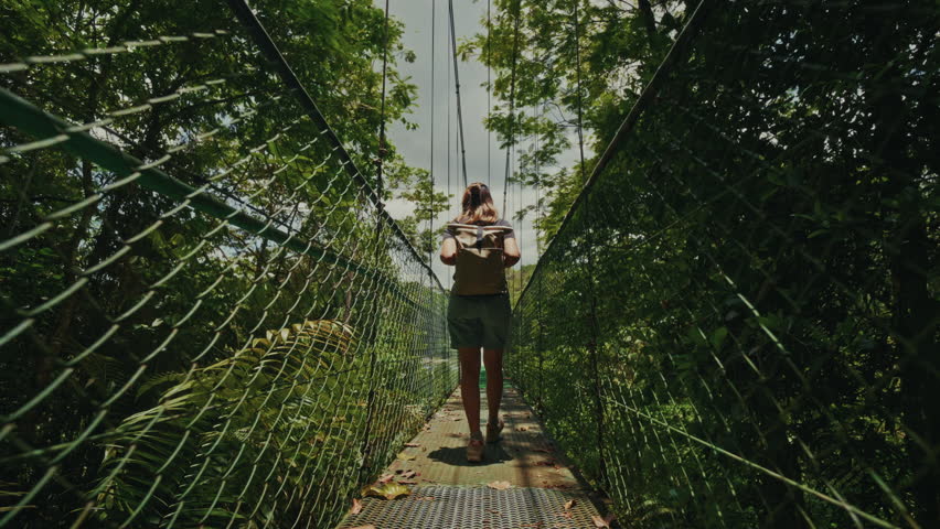 backshot of a young female traveler with a backpack walking on the hanging bridge in Costa Rica enjoying the stunning view of the rainforest.
