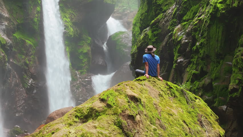 A young boy sits on a mossy rock, captivated by the beauty of a cascading waterfall in Costa Rica, embracing serenity and feeling profound peace and joy as he watches the water flow