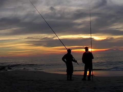 Sunset on the beach with clouds, blue sky and copy space. Two fisherman in the foreground standing with fishing rods