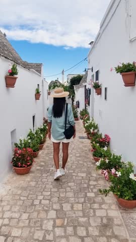 A wanderer meanders along a picturesque cobblestone path, surrounded by vibrant flowerpots. The whitewashed walls create a serene atmosphere under the azure sky, Alberobello Puglia Italy