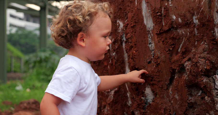Young child stands next to dirt-covered excavator bucket at construction site, almost done exploring this fascinating object. With one finger, he touches the dry clay on heavy equipment, looks at it..