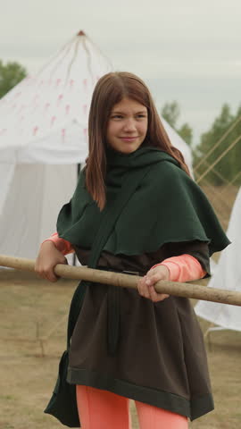 Smiling teenage girl and young man train to fight on spears near white tents. Medieval youth improve fighting skills in preparation for military campaign