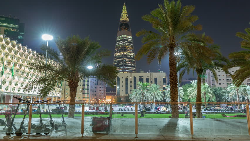 Perspective night timelapse of the King Fahad National Library's illuminated facade with palms and glowing tower in view. People walk across the vibrant square in downtown Riyadh, Saudi Arabia.