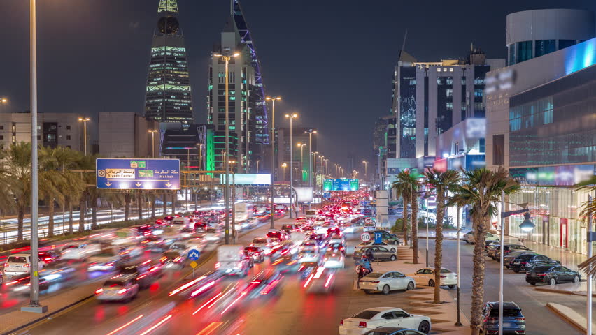 Cityscape aerial night timelapse of Riyadh, Saudi Arabia, featuring illuminated towers, skyscrapers, King Fahd Road and busy traffic. Palms and streetlights line the main highway through the city.