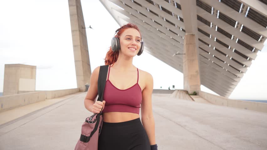 Sporty woman. Young athlete carrying sports bag while confidently walking and listening to music, embodying a healthy, active lifestyle driven by discipline and determination. Girl going to the gym
