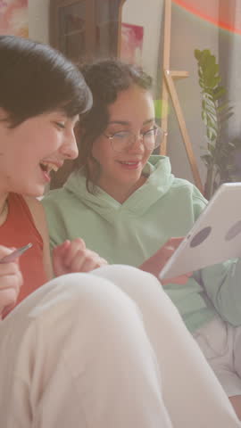 Vertical shot of two teen girls in cozy, sun-drenched room, sitting side-by-side on floor, looking at digital tablet, having conversation and enthusiastically clapping
