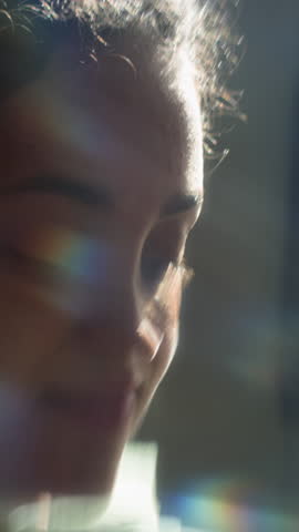 Vertical close-up shot capturing teen girl calm expression as she gazing upwards, her face bathing in soft lighting, with rainbow-like streaks shining on her face with blurred background