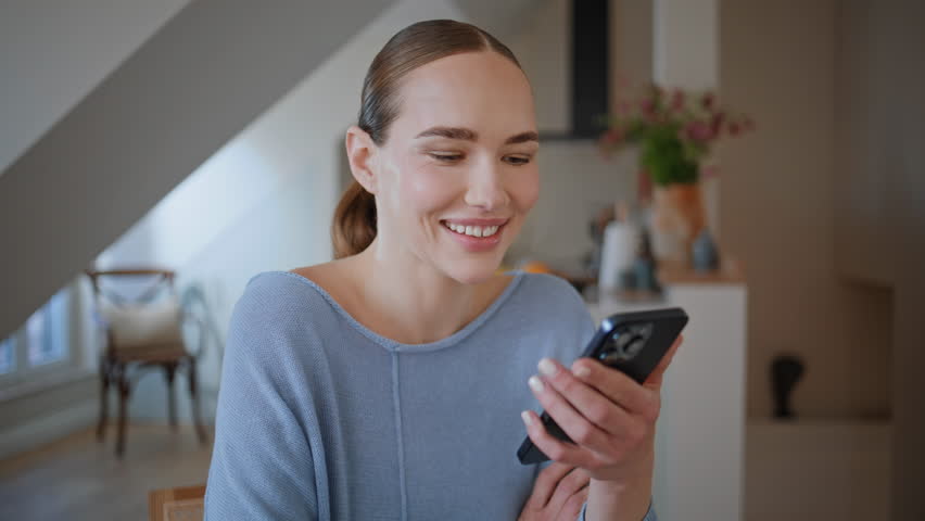 Relaxed woman browsing cellphone in kitchen early morning closeup. Carefree girl looking smartphone screen sitting at home. Calm lady holding mobile phone in white interior enjoying home tranquility 