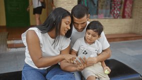 Latin family sitting outdoors in city showing mobile phone to curious child, expressing love and togetherness on an urban street - Powered by Shutterstock - Get 15% off with code: PIKWIZARD15