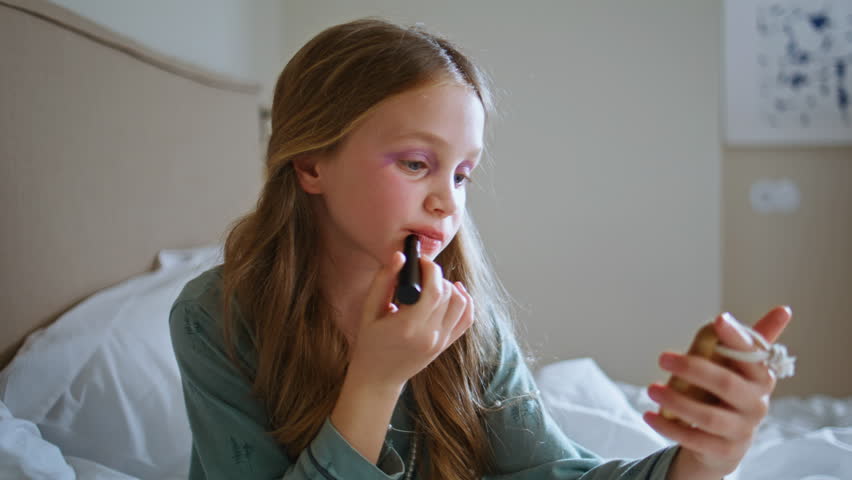 Small girl using makeup in her mothers room closeup. Little longhaired kid putting lipstick imitating adults at home interior. Cute child having fun playing alone at light bedroom. Childhood concept 