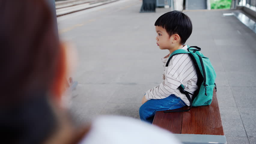 Asian Woman and her Smiling Little boy with a green backpack waiting for a commuter train at the platform in train station.