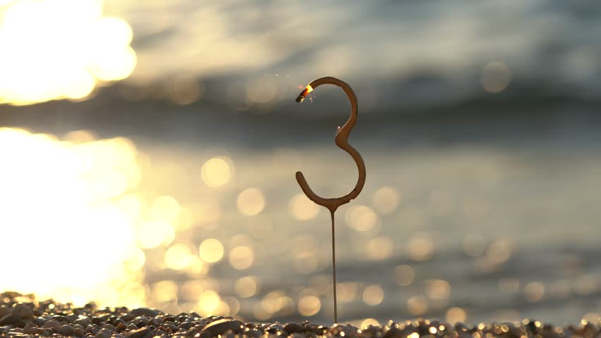 Burning number 3 stick bengal fire on background of sunset waves on sea on sand on beach. Concept of celebrating birthday, 3rd anniversary of business, company. Success, prosperity, congratulations