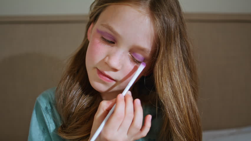 Cute child using makeup in her mothers room closeup. Little longhaired kid putting eyeshadows with brush imitating adults at home. Small girl having fun playing alone at bedroom. Childhood concept 