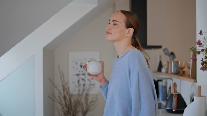 Morning student drinking tea at home kitchen closeup. Smiling attractive woman looking attic window sipping coffee at living room interior. Happy carefree female model enjoying hot cappuccino alone 