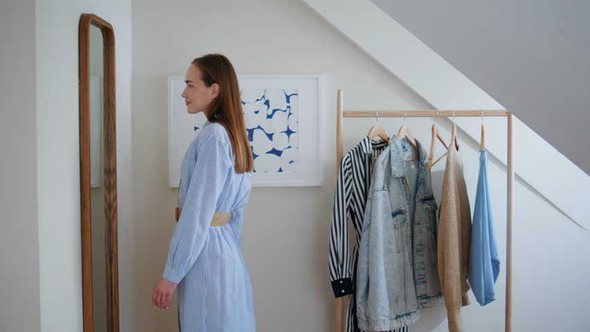 Smiling woman looking mirror reflection choosing outfit for workday or date at home. Happy girl standing in dressing room alone. Positive millennial lady trying on clothes planning look for weekend 