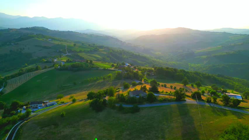 Splendid aerial pullback and pedestal up shot from Monte San Martino with the golden hour sunlight torching through the hills, a lone car passing on the road, and the mighty Sibillini Mountains behind