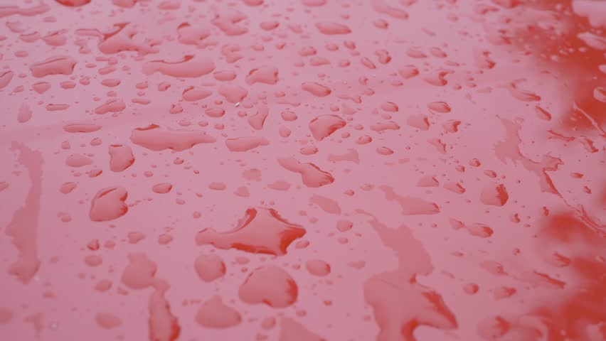 Raindrops scattered across smooth red iron surface, creating small pools of water. Reflections shimmer in tiny droplets as they rest on glossy texture. Weather is rainy