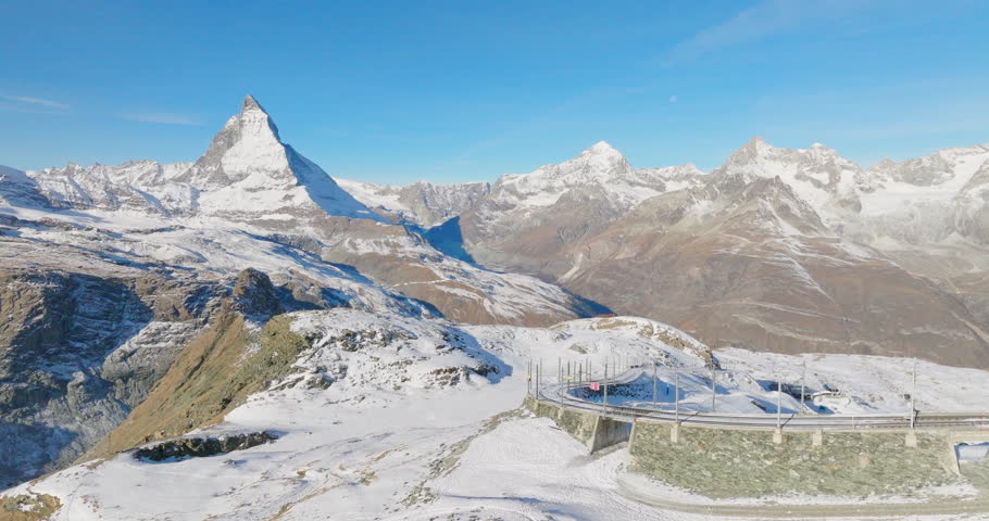 Aerial drone footage of Gornergrat bahn railway running towards summit station with Matterhorn mountain peak background in Zermatt on a sunny winter day. Swiss Alps, Switzerland travel journey trip.