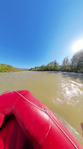 Vertical video. A side-angle shot of a group of rafters paddling down a wide river, wearing yellow helmets and life vests, with trees along the riverbank and a bright sun shining overhead.
