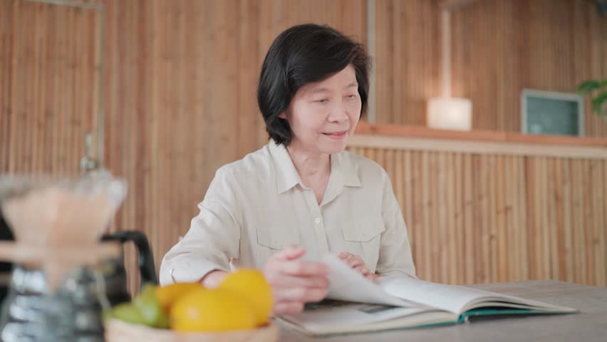 Asian senior woman sitting calmly and relaxed reading a book on a chair with a table, flipping through pages of a book, bamboo house as a backdrop, searching for information, self-improvement concept
