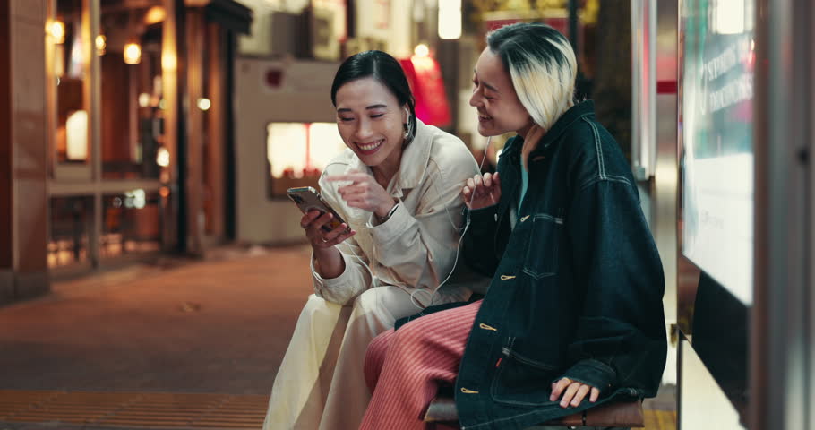 Women, friends and phone at night in city with earphones, laughing and sharing funny video in street. Girl, people and smartphone on urban sidewalk with mobile app, meme or social network in Japan