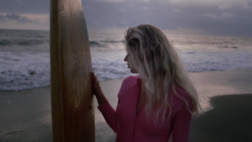 Portrait: beautiful girl with a surfboard. Face of a sexy woman on the shore or beach. Blonde female enjoys the tropical coast of Bali island. Surf board and oink swim suit. Blue hour and sunset
