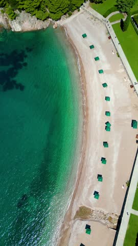 Vertical top view of Sveti Stefan Beach. Famous travel destination in Montenegro. Static footage. Empty beach- Green sun umbrellas. Tourqoise calm water.