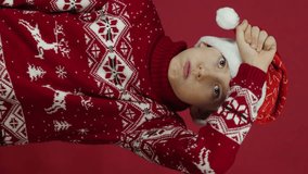 tired frustrated boy 10 years old in christmas sweater and santa hat looking at camera, red studio background, vertical video - Powered by Shutterstock - Get 15% off with code: PIKWIZARD15