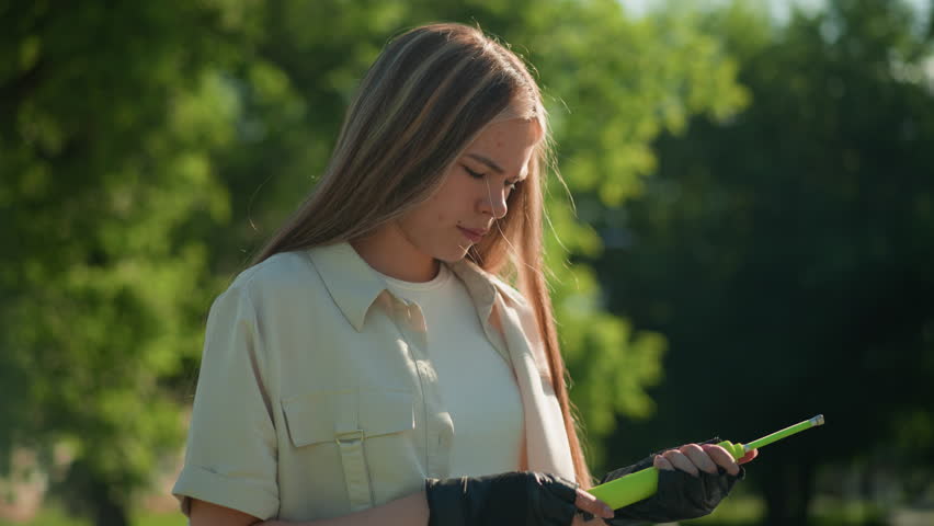 Young woman with blonde hair intently examining green air pump and nozzle in her gloved hands, standing against a backdrop of lush trees and greenery