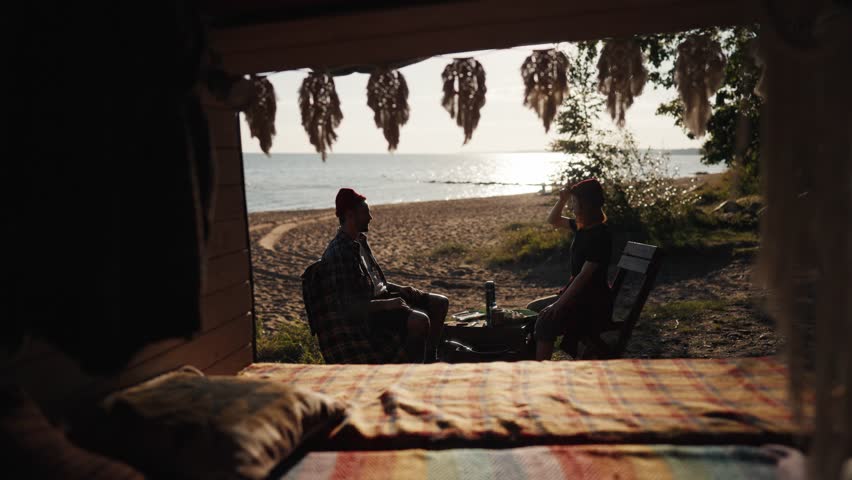 Trip by motorhome, calm breakfast on beach of sea or lake in sunny morning. Two campers relaxing and chatting, enjoying solitude, view from cozy camper car, traveling in nature by auto across country