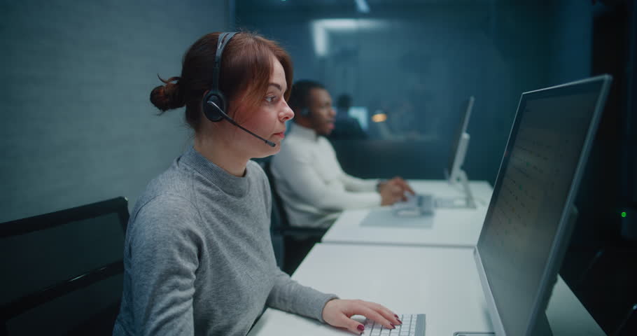 Call center office: Diverse team of technical support specialists working at night. Caucasian female hotline operator talks on headset with customer, works on computer, provides online assistance.