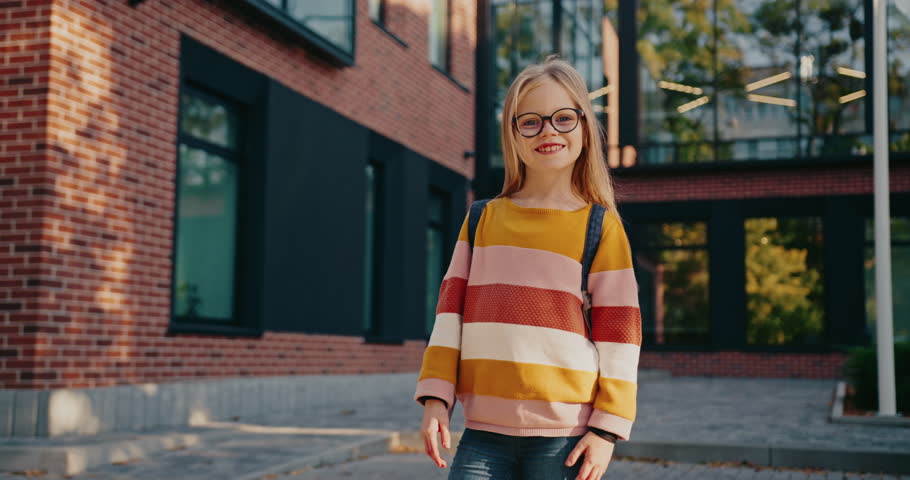 Close portrait of cute Caucasian girl with beautiful blue eyes and blond hair. Wearing glasses. Standing in front of modern school with big windows in background. Ready for school.