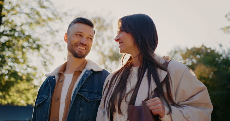 Camera moving around cute Caucasian couple. People looking at each other while smiling. Green trees in blurred background. Bright sundown above tree line. Wearing jacket. Glancing directly.
