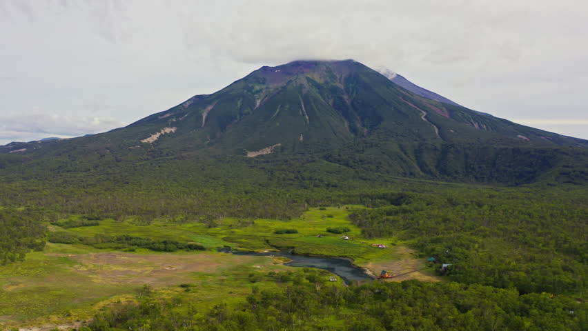 An aerial view of a majestic volcanic mountain surrounded by dense green forests and open meadows. A small river and wooden cabins are visible, creating a peaceful and remote atmosphere ideal for natu