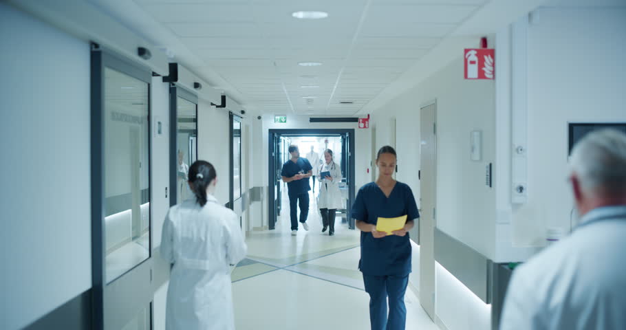 Medical Staff Navigating a Busy Hospital Hallway. Caucasian Male Doctor and Female Nurse Having a Discussion About an Upcoming Surgery and Medical Procedures as They Walk in Corridor with Other People
