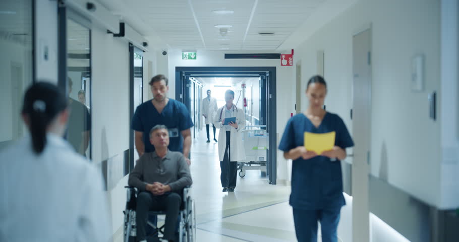 Busy Hospital Hallway Filled with Medical Experts in White Coats and Blue Uniforms. Middle Aged Female Doctor Reviewing a Patient's History on a Tablet Computer as She Heads to the Examination Room