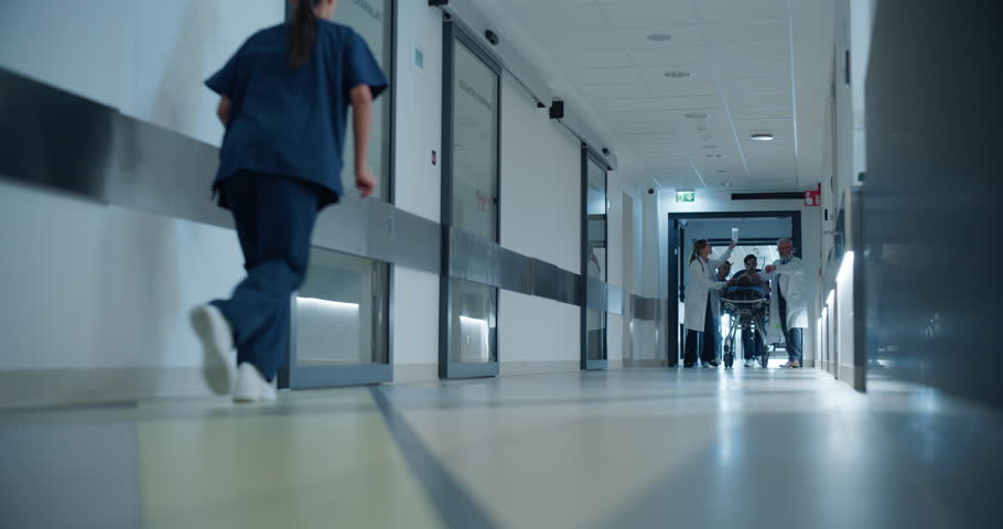Urgent Scene in a Modern Clinic as Paramedics and Doctors Wheel an Unconscious Patient to the Intensive Care Unit. Young Female Nurse Runs Down the Corridor To Help the Team. Low Angle Footage