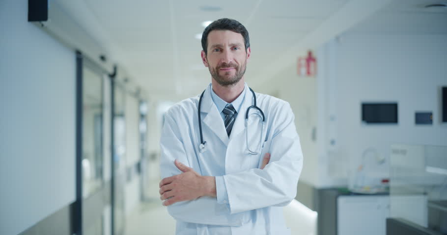 Confident Caucasian Male Doctor in a Crisp White Coat Standing in a Well-Lit Hospital Hallway, Smiling Warmly for the Camera, Showing Professionalism and Approachability of a Medical Worker