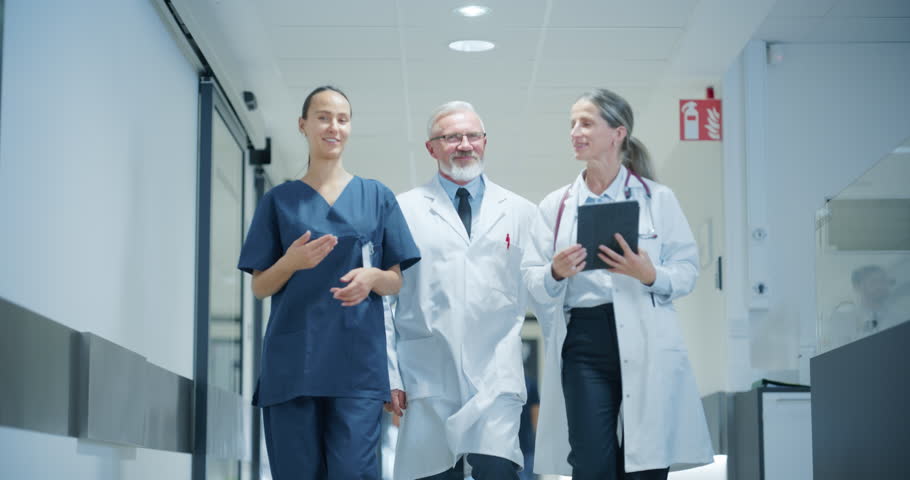 Cheerful Healthcare Team Moving Through a Spacious Hospital Corridor, Preparing for Their Next Shift while Having a Casual Conversation. Female Doctor, Surgeon and Senior Male Working in a Clinic