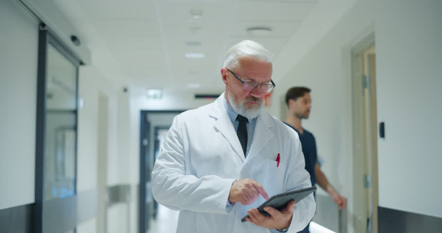 Portrait of a Senior General Practitioner Using Tablet Computer to as He is Walking and Browsing Patient's Medical History. Hospital Personnel Walking Through a Modern Clinic Corridor