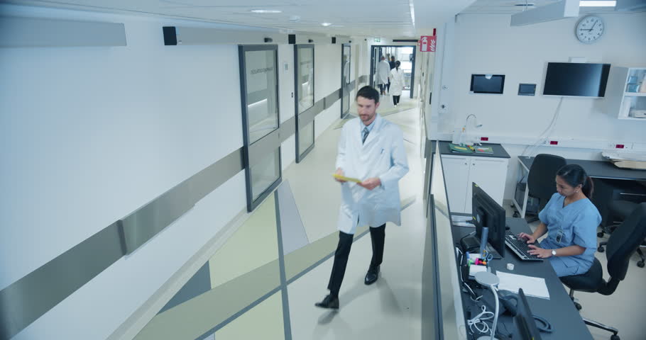 High Angle CCTV Footage with a Busy Hallway with Reception Desk of a Hospital Building, Diverse Female and Male Staff Members Walking, Working. Professional Nurses, Doctors and Personnel at Work