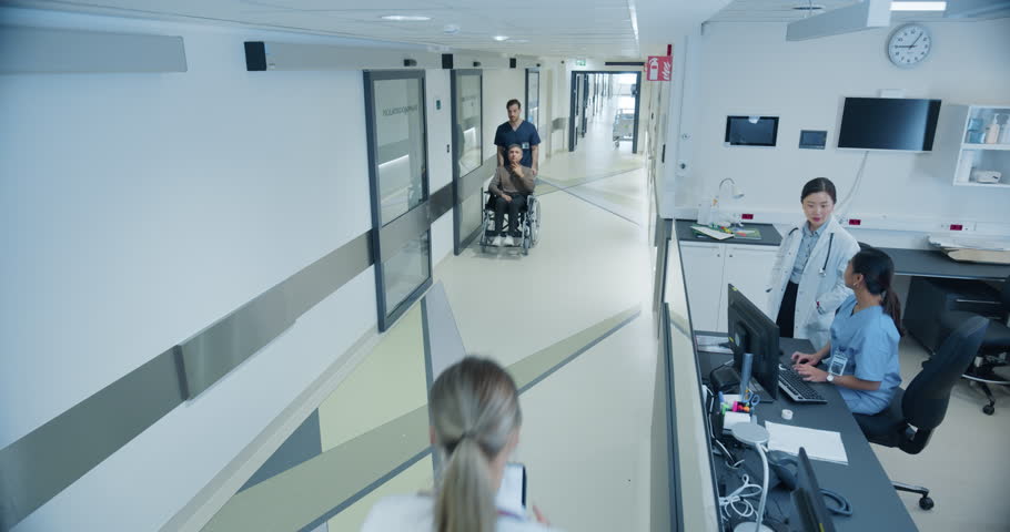 Male and Female Medical Professionals Navigate a Hospital Corridor and Registration Area, Having Healthcare Conversations. Male Nurse Assists a Patient in a Wheelchair. High Angle Security Camera Shot