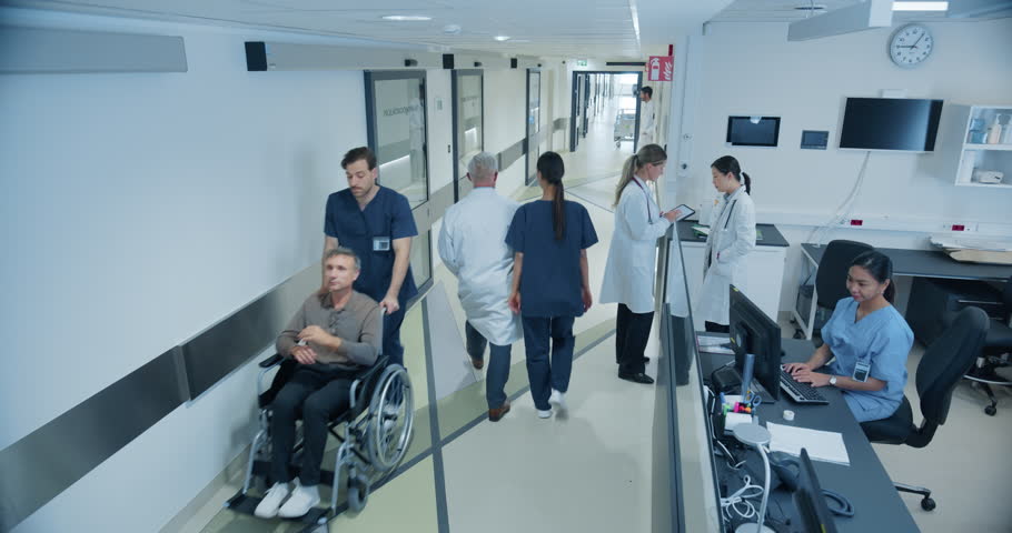 Male and Female Medical Professionals Navigate a Hospital Corridor and Registration Area, Having Healthcare Conversations. Male Nurse Assists a Patient in a Wheelchair. High Angle Security Camera Shot