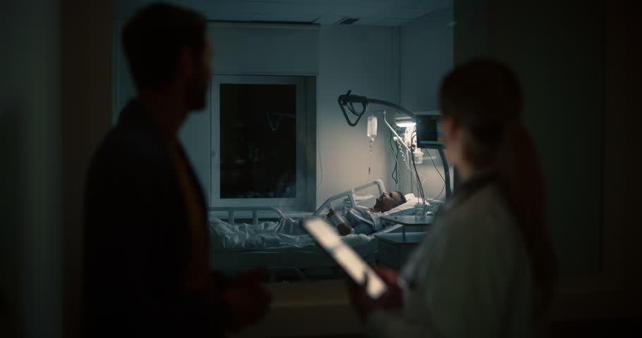 Female Doctor Standing Outside the Patient Ward in a Dark Room, Using a Tablet Computer to Explain Medical Updates to a Concerned Visitor. Nurse Attending to the Patient in the Background
