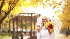 A blonde girl in a white skirt and sweater drinks coffee while standing on the alley of an autumn park with a bouquet of maple leaves and a transparent umbrella - Powered by Shutterstock - Get 15% off with code: PIKWIZARD15
