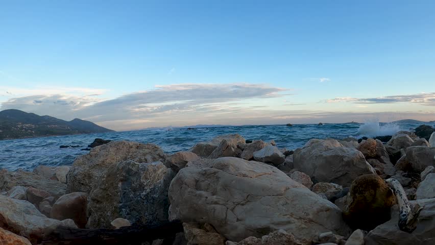 A rocky shoreline with clear blue water and distant mountains under a partly cloudy sky. The waves crash gently against the rocks, creating a serene coastal scene during the late afternoon.